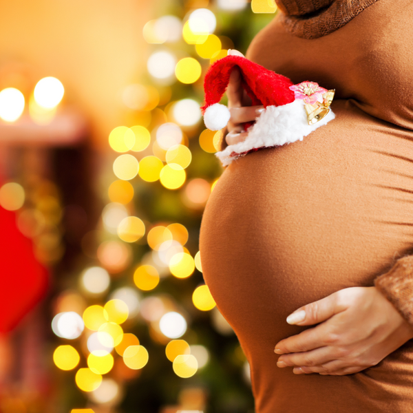 Close-up of a pregnant belly with a hand holding a Santa hat, Christmas tree lights in the background