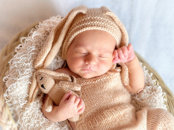 Sleeping newborn in a knitted bunny bonnet cuddling a knitted bunny toy in a basket