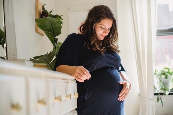 Pregnant woman leaning against a dresser and looking down at her belly with a hand resting on it