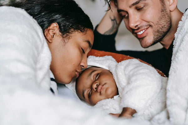 Two parents leaning in to kiss their sleeping newborn wrapped in a white blanket