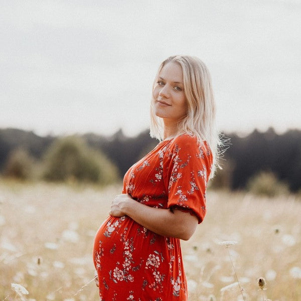 Pregnant woman in a red floral dress smiling over her shoulder while standing in a meadow