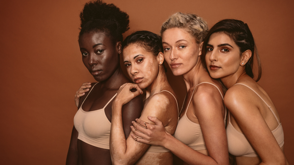 Four women in nude-toned underwear posing together against a warm brown background