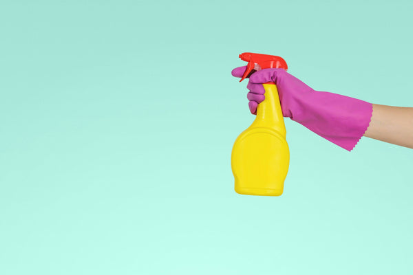 Gloved hand holding a yellow spray bottle with a red trigger against a teal background