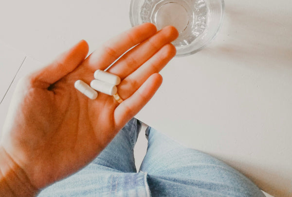 Hand holding white supplement capsules with a glass of water nearby