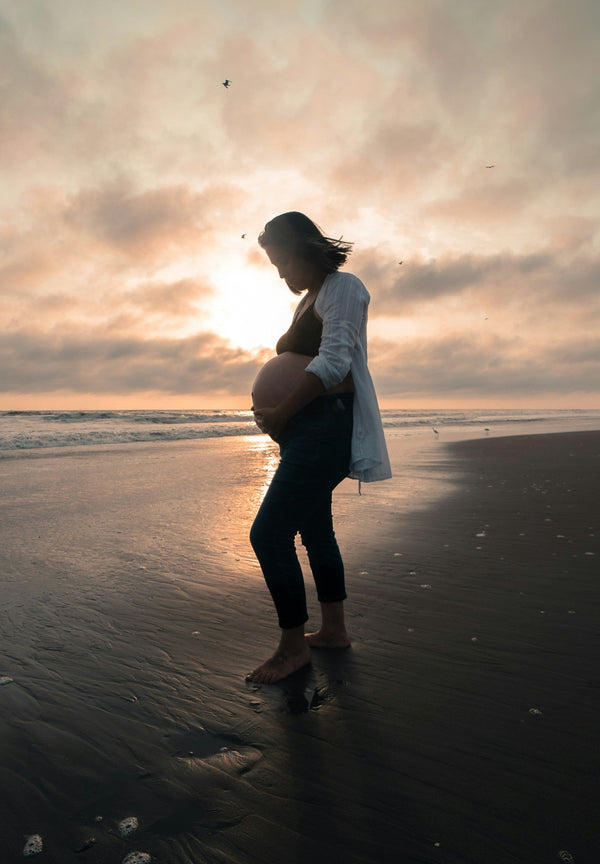 Silhouette of a pregnant woman standing on a beach at sunset holding her belly