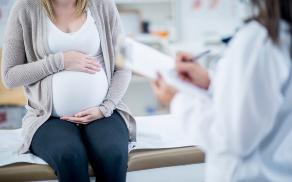 Pregnant woman sitting on an exam table with her hands on her belly while a doctor writes notes