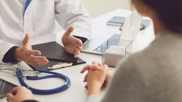 Doctor gesturing while talking to a patient across a desk with a stethoscope and clipboard