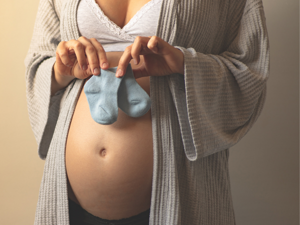 Close-up of a pregnant woman in a robe holding a pair of blue baby socks against her belly