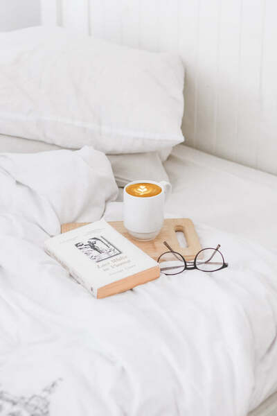 Cup of coffee, a book and glasses resting on a tray on a white bed