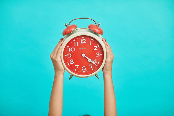 Hands holding up a red alarm clock against a teal background