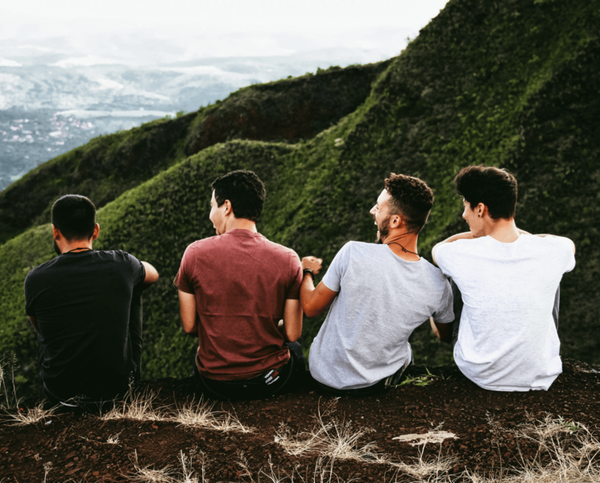 Four men sitting on a hillside overlooking a green valley, backs to camera