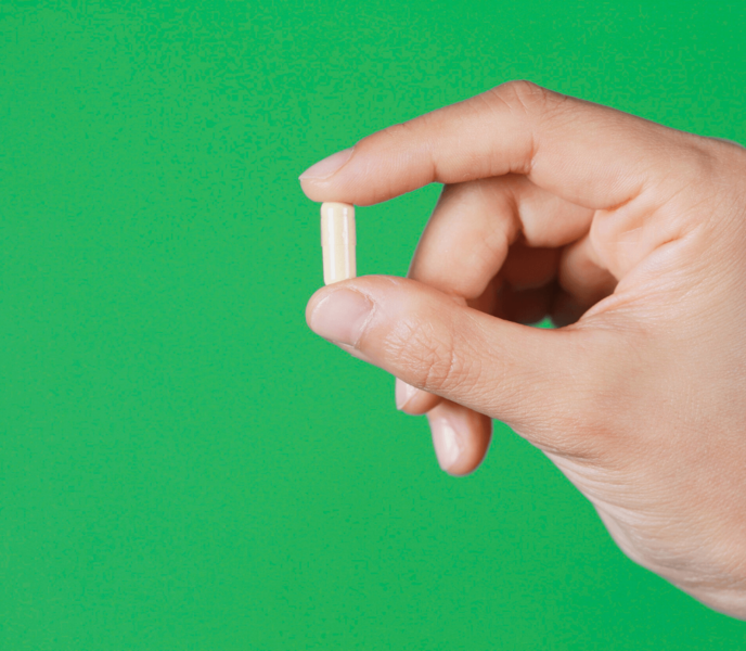 Hand holding a single beige capsule against a green background