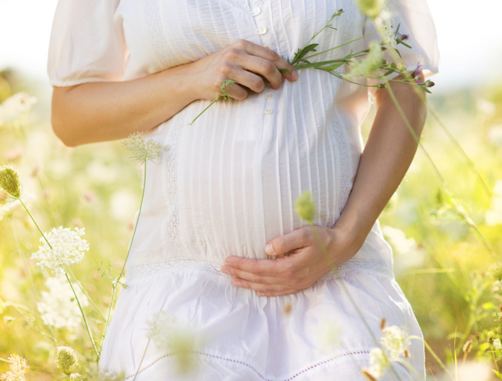 Pregnant woman in a white dress cradling her bump while holding wildflowers in a sunny meadow