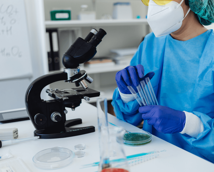 Lab researcher in blue scrubs and gloves holding test tubes beside a microscope