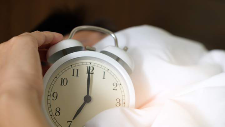 Hand reaching to silence a white alarm clock beside a person in bed