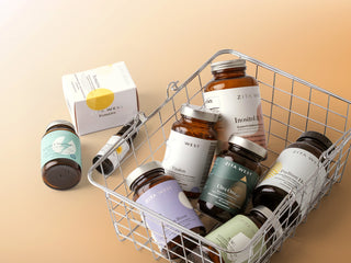 A styled product shot of multiple supplement bottles and a box from ZITA WEST arranged in and around a metal wire basket on a warm beige background.