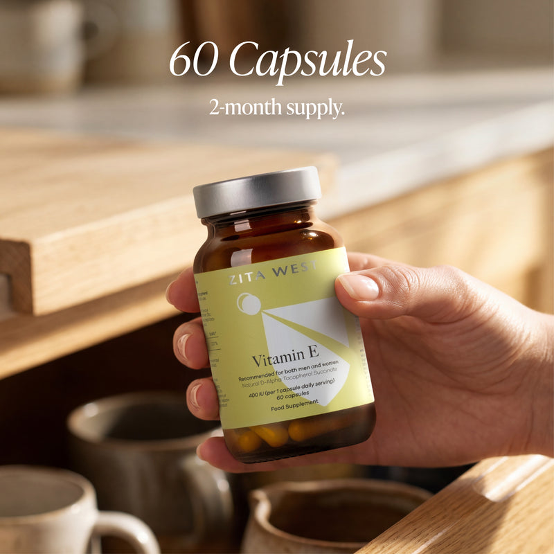 Hand holding a bottle of Zita West Vitamin E capsules with a kitchen background.