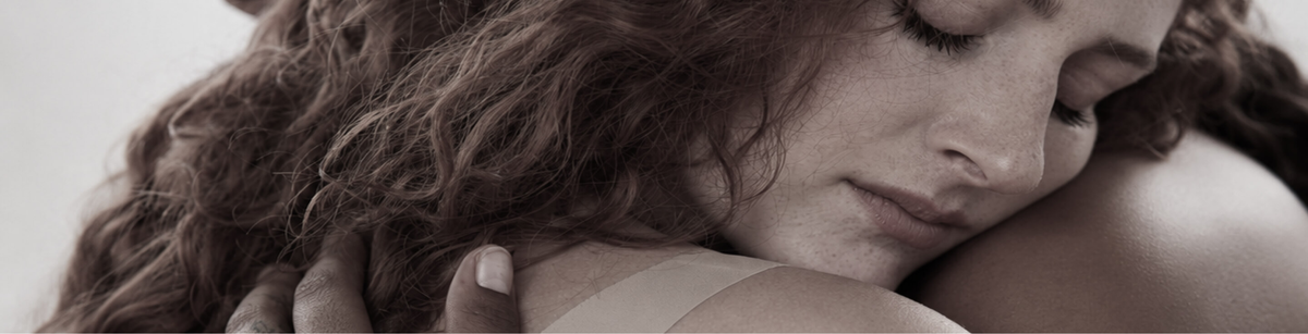 A close-up lifestyle shot of a woman with long, wavy brown hair resting her head on another person's shoulder, captured in a soft, warm sepia-toned photograph.